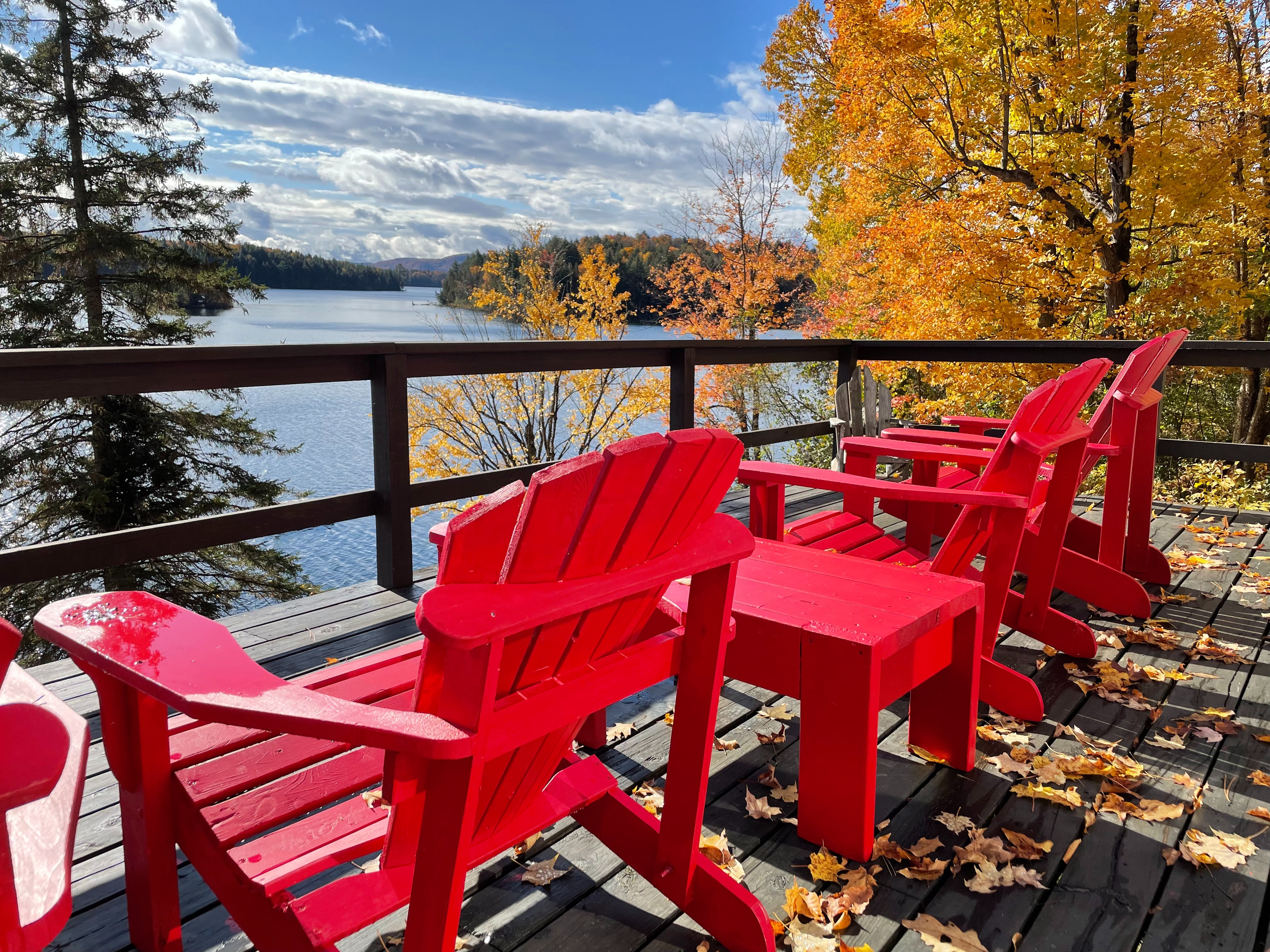 Three matching red chairs on a wooden deck overlooking a lake, surrounded by trees with autumnal yellow leaves.