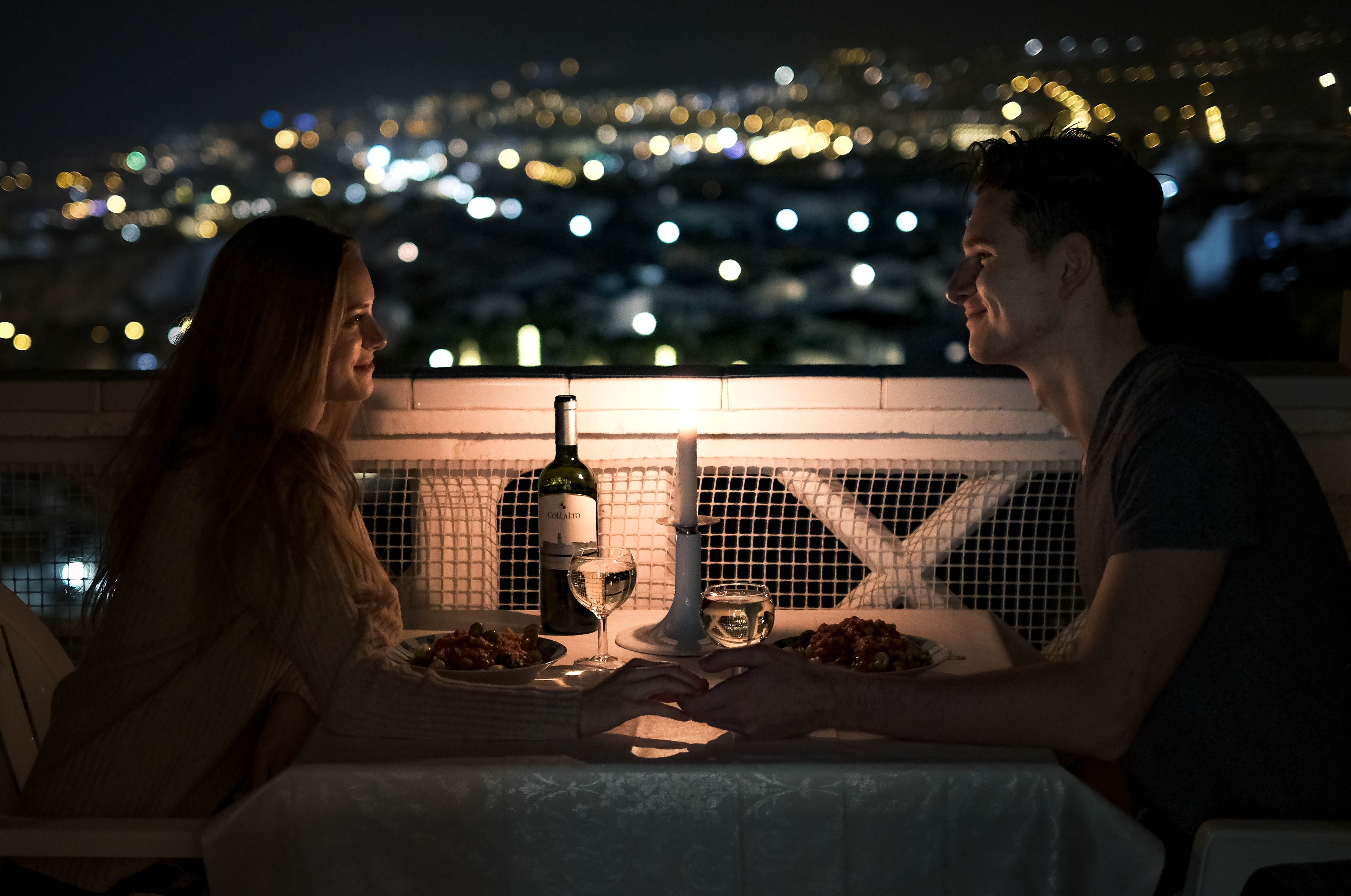 Two people enjoying a candle-lit meal on a terrace with a blurred lit-up city in the background.